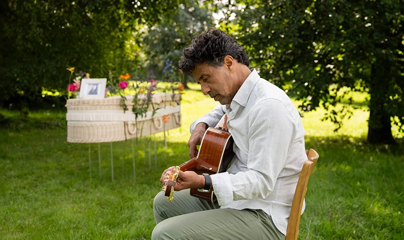 Man speelt gitaar naast rotan uitvaartmand