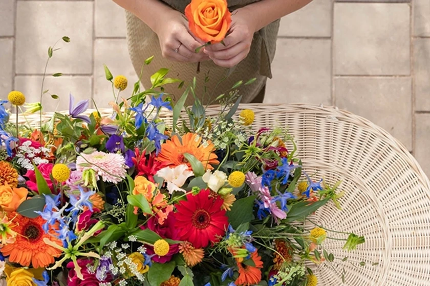 Vrouwlijke handen met roos naast uitvaartmand met kleurrijk veldbloemen rouwstuk