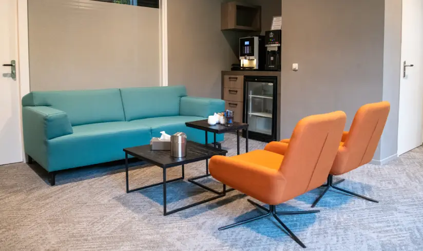 Kamer met een blauwe bank en 2 oranje fauteuils, en een koelkast en koffiezetapparaat in de hoek.