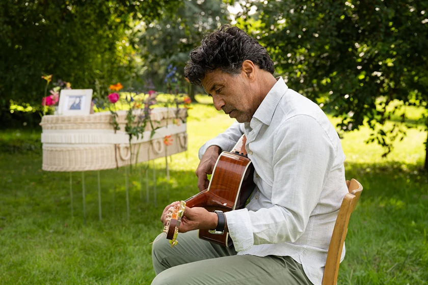 Man speelt gitaar naast kleurrijke uitvaartmand in de groene natuur