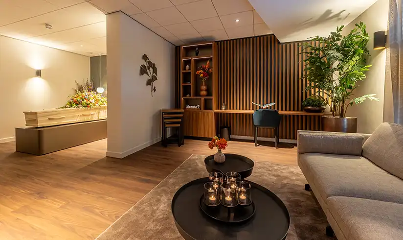Een kamer met een uitvaartkist met bloemen en een zithoek in een aangrenzende kamer