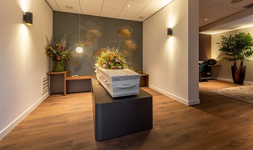 Een kamer met een tafel en stoelen en een uitvaartkist met bloemen in een aangrenzende kamer