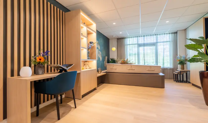 Rouwkamer met een bureau met stoel en de kist met bloemen erbovenop