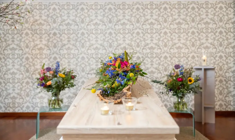 Een ruimte met een houten uitvaartkist met bloemen erbovenop en aan het hoofd