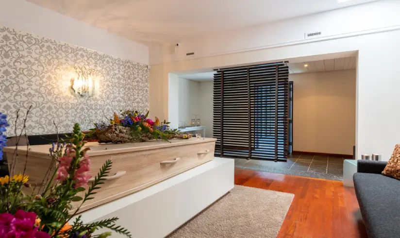 Een kamer met een uitvaartkist met bloemen en afscheiding van de hal