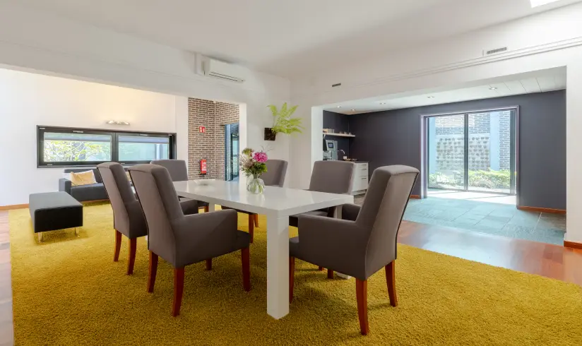 Grote ruimte met een witte tafel en 6 grijze stoelen en een geel tapijt op de vloer