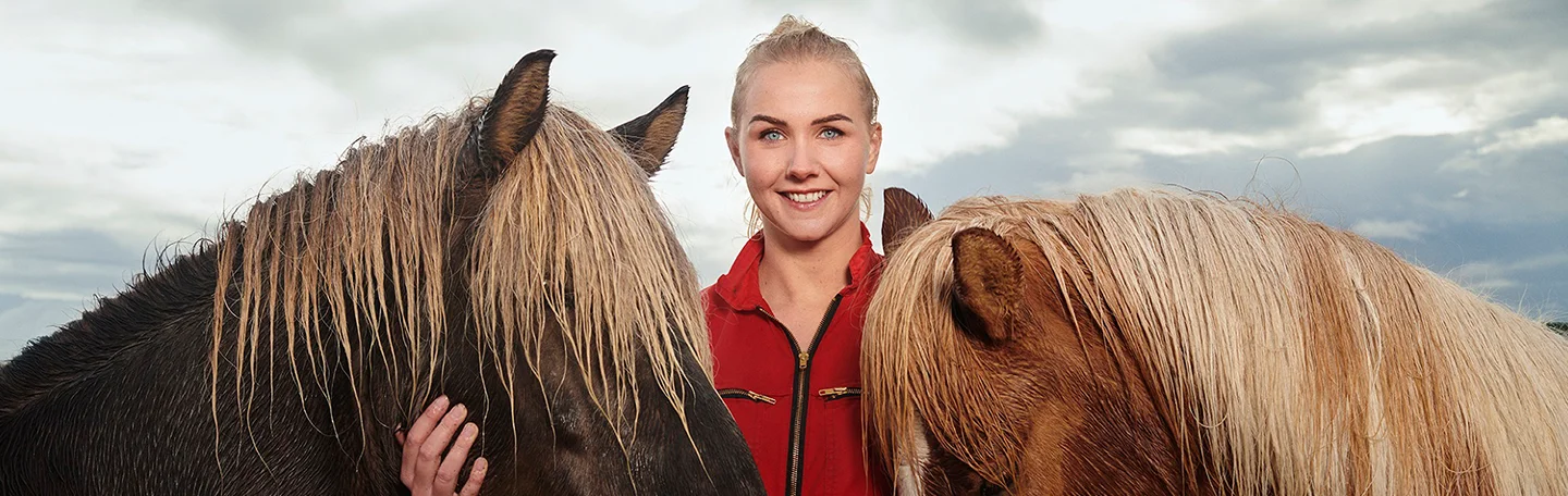 Blonde vrouw in rood overall staat in weiland tussen twee paarden