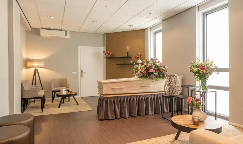 Een kamer met een uitvaartkist met bloemen en een zithoek met 2 stoelen