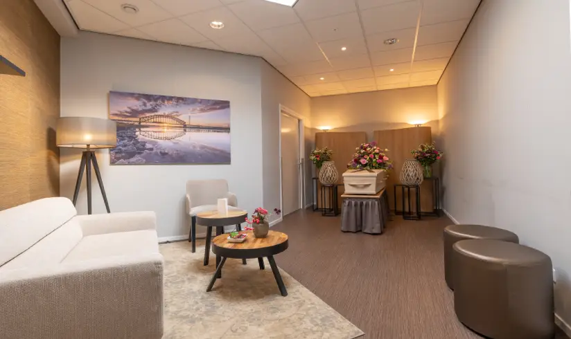 Kamer met een bank, een stoel, en 2 poefen en achterin een uitvaartkist met bloemen