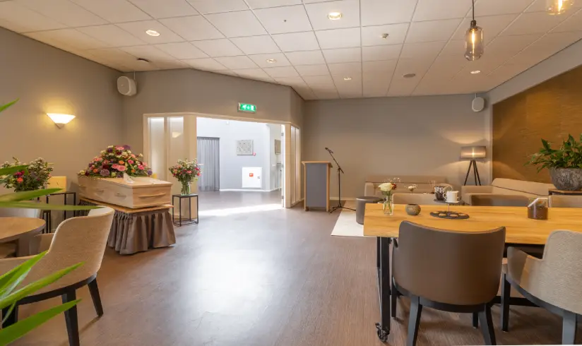 Grote ruimte met tafels en stoelen en een houten uitvaartkist met bloemen