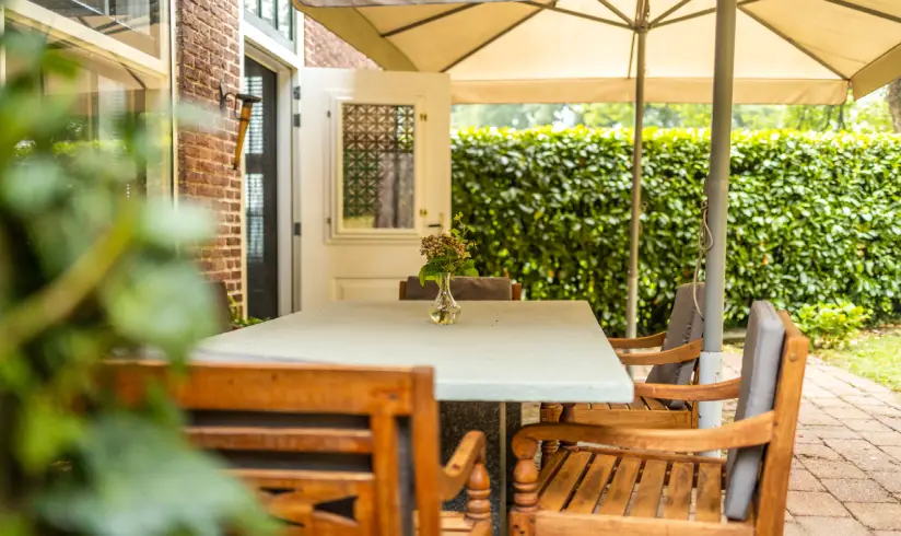 Onder een lichtgekleurde parasol staat een tafel met houten stoelen eromheen