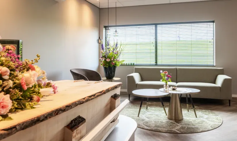 Kamer met een houten uitvaartkist met bloemen en een zithoek met een bank en een stoel