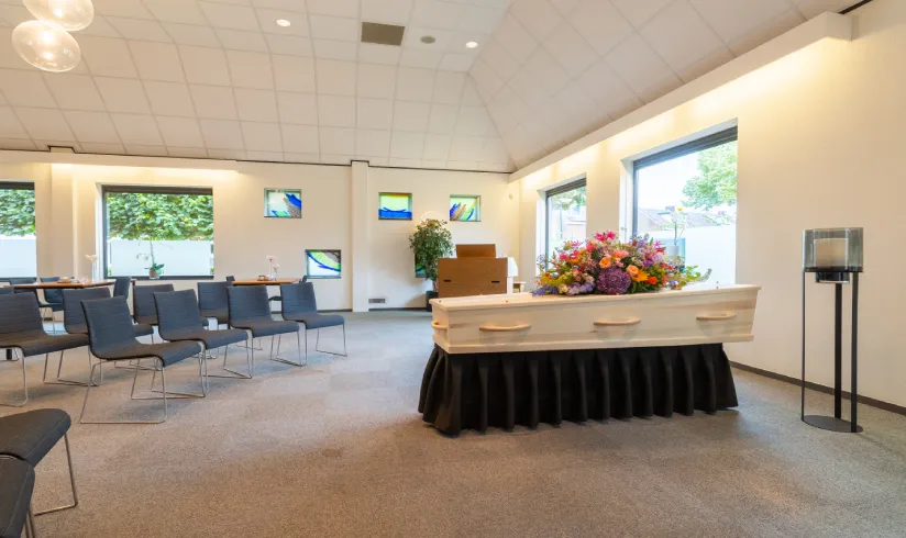 Grote ruimte met rijen stoelen en een uitvaartkist met bloemen