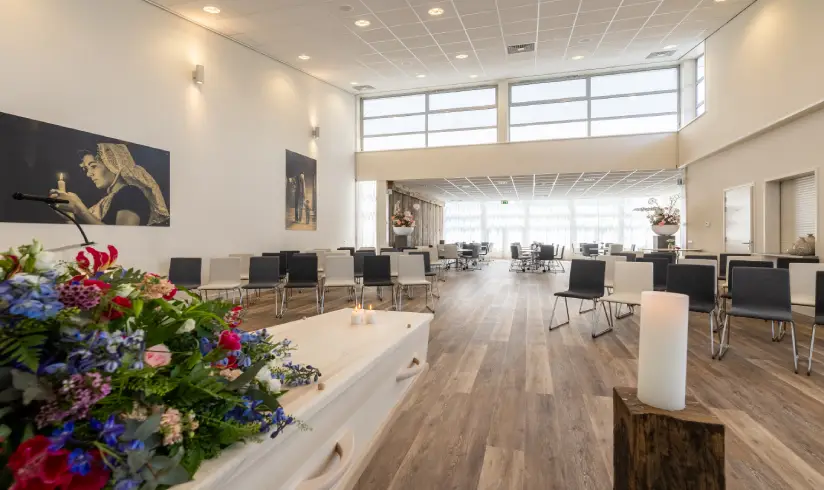 Grote ruimte met een uitvaarkist met bloemen en rijen zwarte en witte stoelen