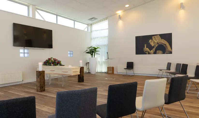 Grote ruimte met een houten uitvaartkist met bloemen en rijen met stoelen