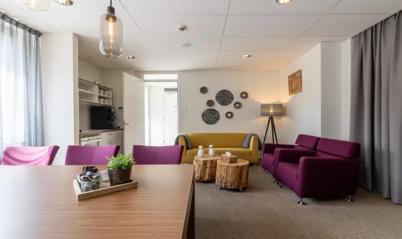 Woonkamer met een tafel en paarse stoelen, een gele bank en 2 paarse fauteuils
