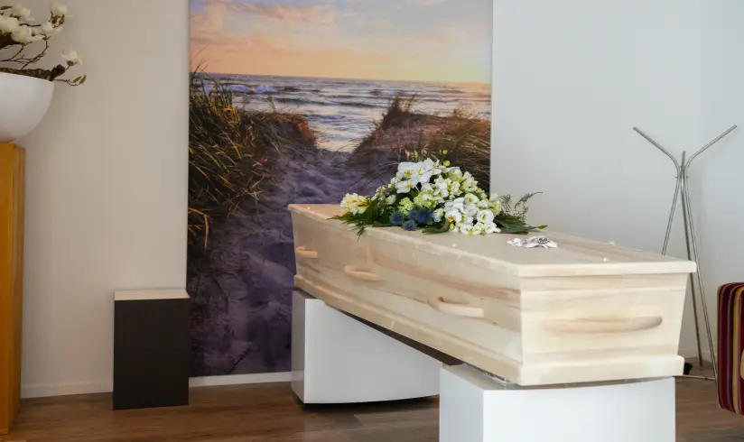 Kamer met een houten uitvaartkist met bloemen en een schilderij van het strand