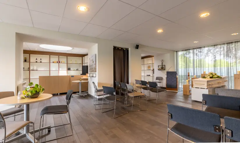 Grote ruimte met een tafel met stoelen, rijen stoelen en een houten uitvaartkist