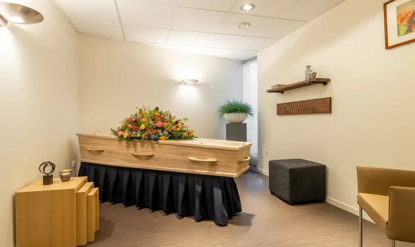 Een kamer met een houte uitvaartkist en een beige stoel naast de muur