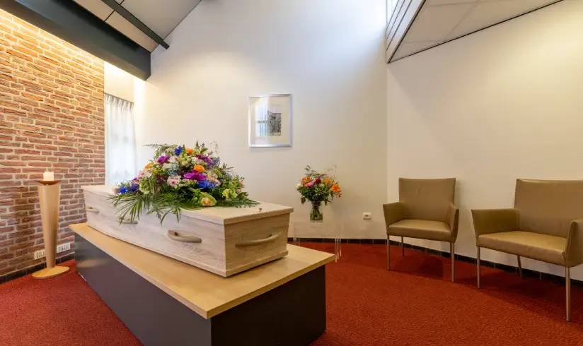 Ruimte met een uitvaartkist met bloemen en 2 stoelen tegen de muur