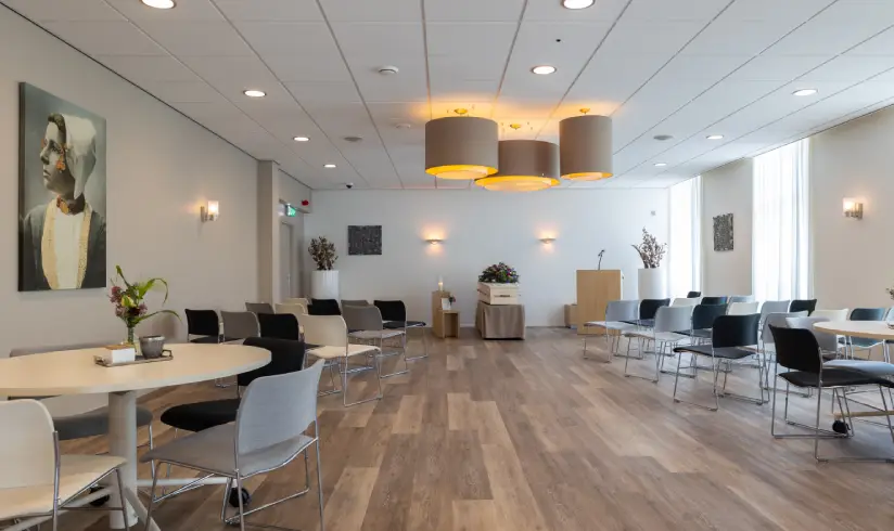 Grote ruimte met tafels en stoelen en achterin een uitvaartkist met bloemen