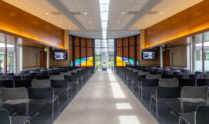 Grote zaal met rijen zwarte stoelen en 2 grote tv schermen voorin aan de muur
