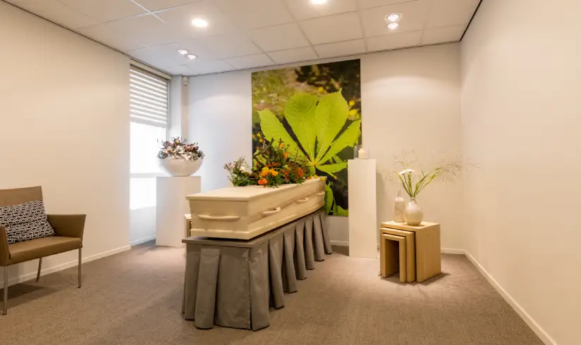 Kamer met een uitvaartkist met bloemen en een stoel aan de muur