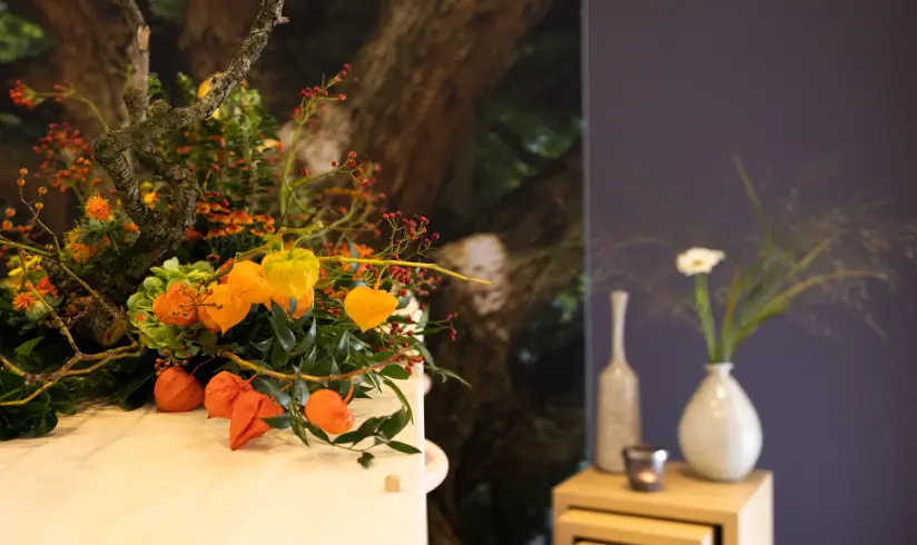 Close up op een uitvaartkist met bloemen en een tafeltje met decoratie op de achtergrond