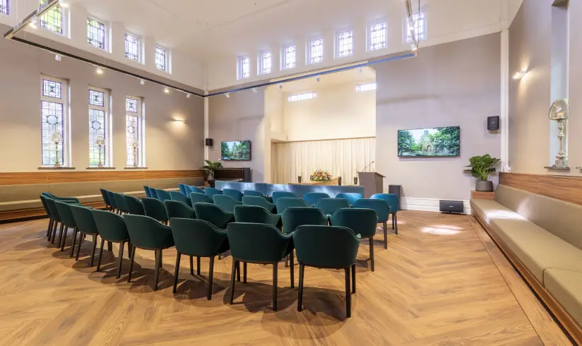 Grote zaal met 4 rijen met blauwe stoelen en vooraan een spreekgestoelte en een houten uitvaartkist