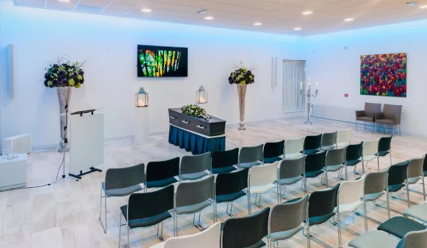 Grote zaal met rijen stoelen en een uitvaartkist met bloemen