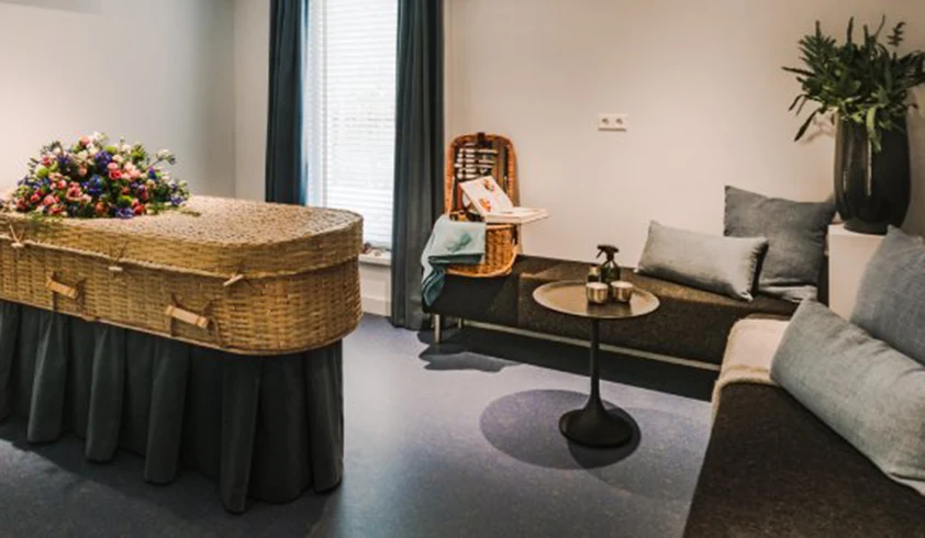 Kamer met zwarte banken met grijze kussentjes en een rieten uitvaartkist met bloemen