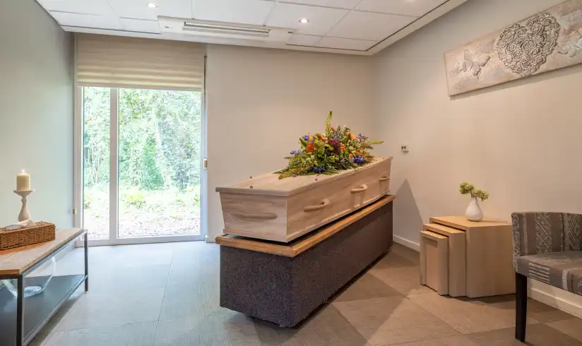 Kamer met een uitvaartkist met bloemen en een tafeltje tegen de muur