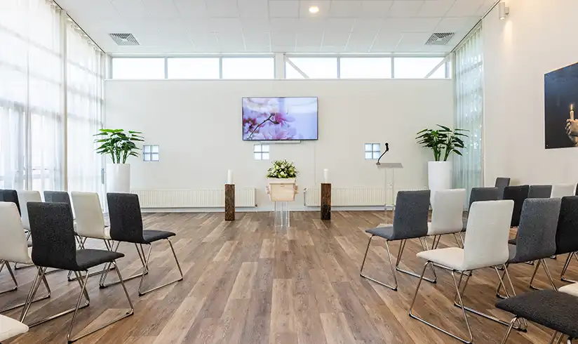 Grote ruimte met een houten uitvaartkist met bloemen en rijen met stoelen